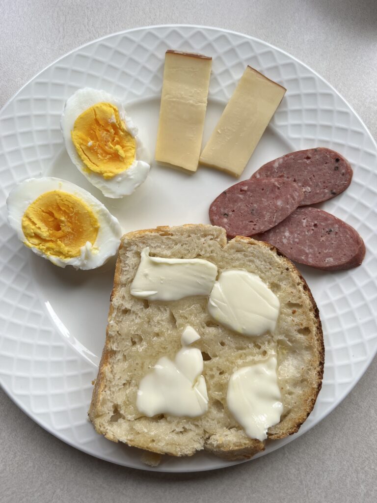 Sourdough sandwich bread with butter on a plate with German sausage, smoked gouda, and boiled egg cut in half. 
