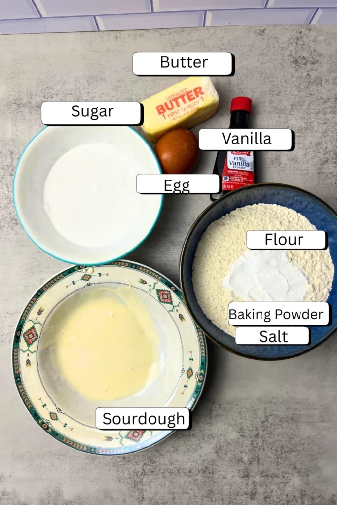 Ingredients for sourdough sugar cookies neatly arranged on a countertop: flour, sugar, butter, egg, vanilla extract, baking powder, salt, and active sourdough starter.