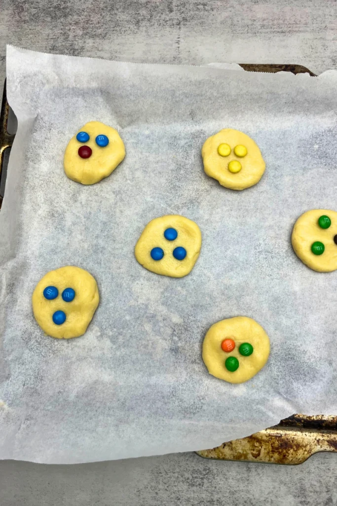 Sourdough sugar cookie dough rolled into small balls, with colorful M&Ms pressed on top, arranged on a baking sheet ready to bake.