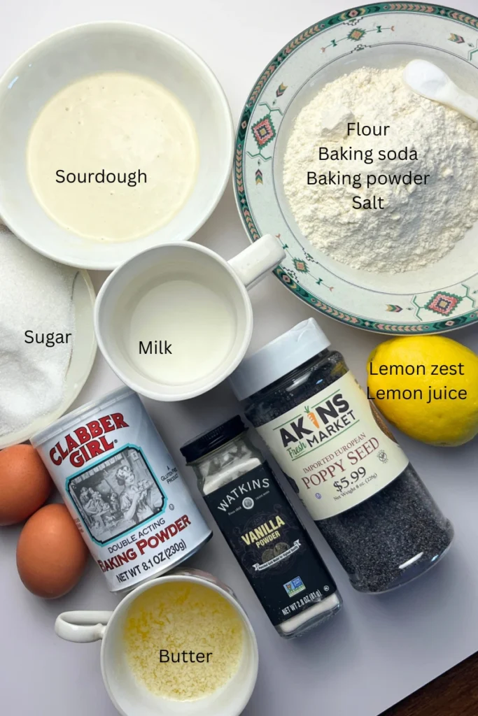 1 Flat lay of sourdough lemon poppy seed muffin ingredients including sourdough discard, flour, sugar, eggs, butter, fresh lemons, milk, baking powder, baking soda, salt, vanilla extract, and poppy seeds.